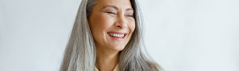 Pretty laughing Asian lady with natural silver hair poses for camera on light grey background in...