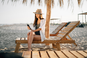 young girl in tropical resort during her vacation sits on wooden deck chair near straw umbrellas on...