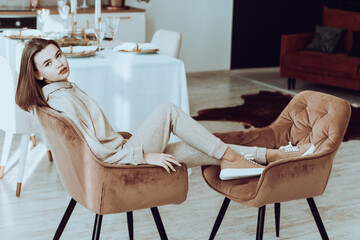 photo of a women in beige sports suit and stylish leather sneakers posing in the chair on a kitchen . selective focus
