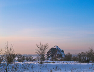 Bulk mountain and trees in a winter field under the rays of the evening sun against the sky