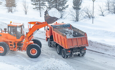 Big orange tractor cleans up snow from the road and loads it into the truck. Cleaning and cleaning of roads in the city from snow in winter. Snow removal after snowfall and blizzards. 
