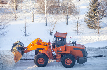 Big orange tractor cleans up snow from the road and loads it into the truck. Cleaning and cleaning of roads in the city from snow in winter. Snow removal after snowfall and blizzards. 