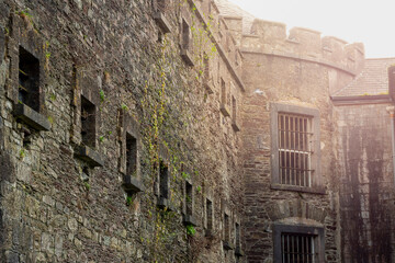 Old stone jail wall with small windows with metal bars. Sun flare. Law correction facility with hard living conditions. Cork Gaol, Ireland. Male and female prison.