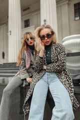 Cool trendy two women girlfriends with fashion sunglasses in stylish clothes with fashionable leopard coat and jeans sits on the street near vintage building