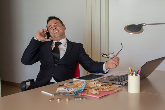 Image of a smiling businessman in his office talking on the phone and bragging about showing his money earned in online finance and investing