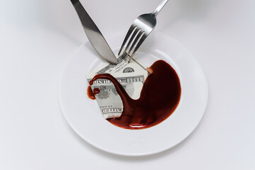 A fork and knife cut through a hundred-dollar bill that lies in a white plate next to blood-like...