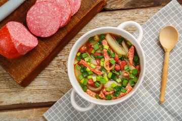 Jewish cuisine bean soup cholit in a tureen on a gray napkin next to a sausage and a flatbread and a wooden spoon.