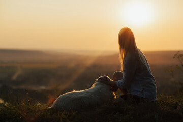 Young woman stroking her dog as they sit on the mountain at beautiful sunset. Warm light from sunset.