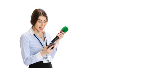 Excited young girl, female journalist holding reporter microphone and looking at phone isolated on white background. Concept of social media, press, news, information