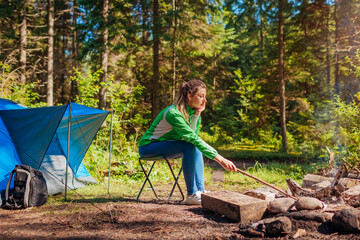Naklejka na ściany i meble Woman relaxing by campfire in forest sitting next to tent. Summer camping. Traveling alone