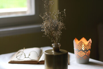 Open book, reading glasses, candle holder with lit candle and vase with gypsophila flowers. Dark background, selective focus.