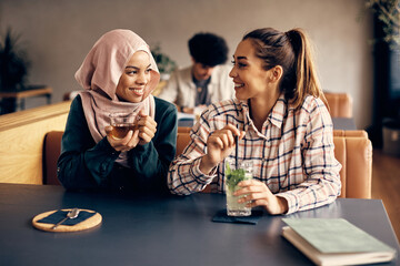 Young happy Muslim woman and her Caucasian female friend talk while relaxing in a cafe.
