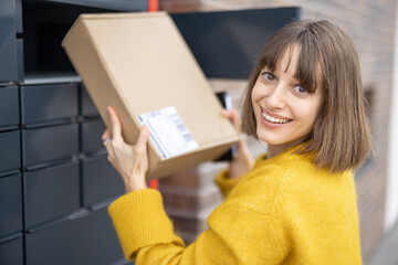 Young woman getting parcel from cell of automatic post terminal outdoors. Concept of contactless and smart delivery. Idea of modern shipping and logistics. Woman wearing yellow sweater