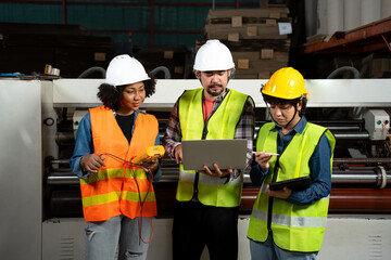 Front view of three factory workers, Asian man and woman, and African woman, in a safety vest and helmet, standing in front of industrial machines looking at data in a laptop held in hand at factory.