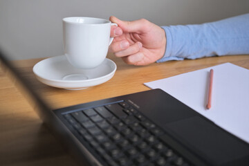 A man at his desk in front of a laptop raises a white cup of coffee to drink, in front of him is a white sheet of paper and a pencil