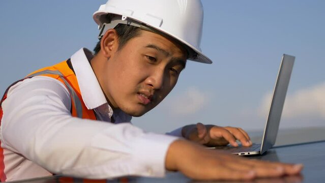 Close up, Young Asian technician man checking operation of sun and cleanliness of photovoltaic solar panel and typing on laptop computer while working in solar farm