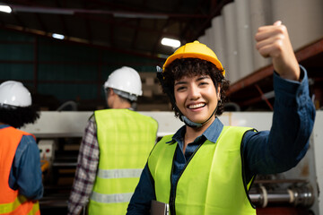 Selective focus, front view of a happy pretty short-haired Asian female factory worker in a vest and helmet, showing thumbs up while smiling at camera with blurred workers working in the background.