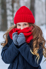 A beautiful teenage girl with brown eyes hides her face in a red snood and smiles. Walk through the forest in the cold season in warm clothes. Knitted warm accessories in actual colors.