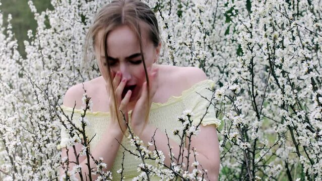 Young woman blowing nose and sneezing in tissue in front of blooming tree. Seasonal allergens affecting people. Beautiful lady has rhinitis.