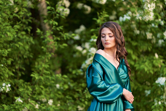 Portrait of a green-eyed beauty in green silk clothes against a background of blooming apple trees. Copy space.
