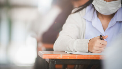 Student taking exam while wearing face mask due to coronavirus emergency. Young woman sitting in...