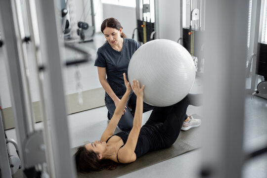 Woman doing exercises with fitness ball with rehabilitation specialist at the gym. Concept of physical therapy for health and recovery. Idea of recovery after pregnancy