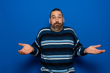 Fototapeta premium Puzzled hesitant bearded fashionable male full of doubts, should decide or choose between two proposals, pouty lips, has indecisive expression, shows uncertainty, gestures against blue background