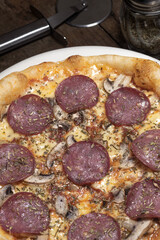 Pizza lista para comer con salsa roja, queso mozzarella, champiñon, salami, pimienta y orégano en un plato blanco sobre un fondo de madera junto a un cortador de pizza
