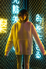 Girl in a white hoodie and leggings stands near a metal mesh with neon symbols.