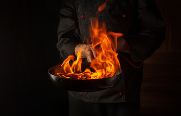 Professional chef cooking food in pan with fire flame on black background. Restaurant and hotel...