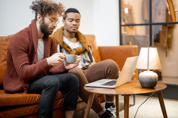 Gay couple with different nationality sitting together on a couch, talking and watching something on laptop at home. Concept of homosexual relations and lifestyle at home