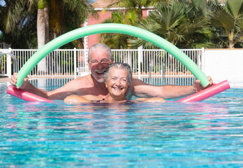 Cheerful adult happy senior couple having fun in outdoors swimming pool doing exercise with swim noodles. Smiling retired people playing together in the pool water under the sun - Powered by Adobe
