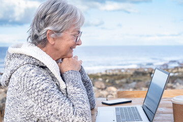 Smiling attractive mature senior adult professional business woman in remote working on laptop computer by the beach wearing eyeglasses, sitting at the wooden table looking at computer screen