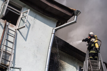 Firefighters on ladders in oxygen masks extinguish the fire in an old house in the middle of the...