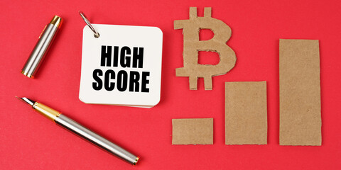 On a red surface lie the bitcoin symbol, a pen and a notepad with the inscription - High Score