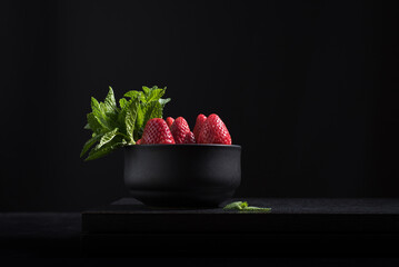 Fresas enteras con hojas de menta en un recipiente negro sobre una mesa de madera negra y fondo oscuro  © R. Guas