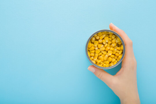 Young Adult Woman Hand Holding Opened Metallic Preserved Can With Yellow Corn On Light Blue Table Background. Pastel Color. Closeup. Empty Place For Text. Top Down View.