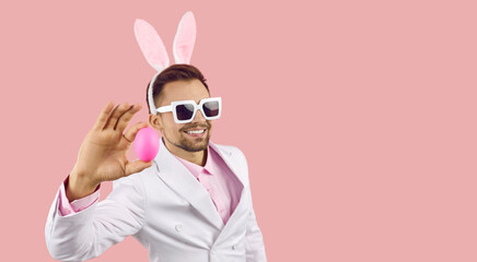 Happy handsome young rabbit man wearing white suit, cool sunglasses and funny bunny ears standing on pink background with copy space on right side, looking at camera, smiling and showing Easter egg