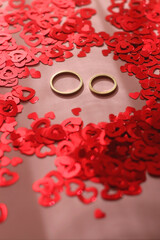 Gold wedding rings and red heart confetti on pastel pink background. Selective focus.