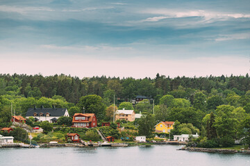 Fototapeta premium Sweden. Many Beautiful Red Swedish Wooden Log Cabins Houses On Rocky Island Coast In Summer Sunny Evening. Lake Or River Landscape.
