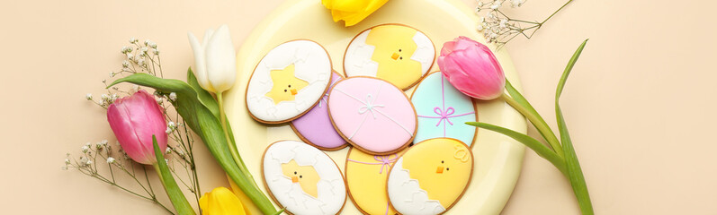 Plate with creative Easter cookies and flowers on beige background