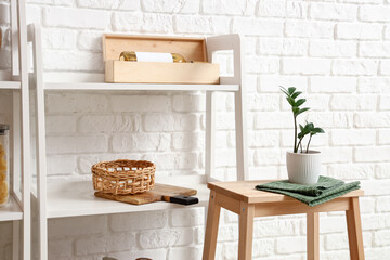 Modern shelf unit with kitchenware and stool near white brick wall