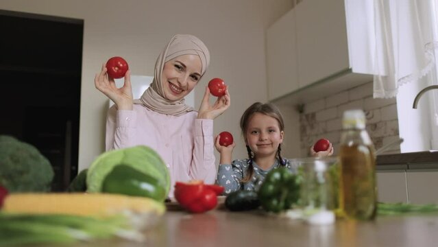 Arabian mother sit at kitchen table with little cute preschooler daughter teach cooking salad while have fun hiding their eyes behind tomatoes. Nanny enjoy weekend preparing lunch food with child