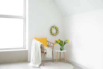 Chair and table with bouquet of tulips near light wall with handmade Easter wreath in room