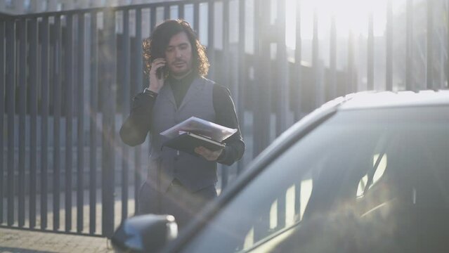 Portrait of confident elegant man standing in sunrays outdoors with phone calling. Positive successful Caucasian manager with luxurious car downtown in the morning. Slow motion