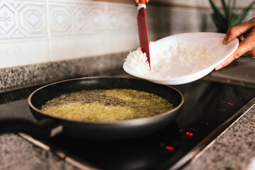 Hands of a young caucasian woman adding chopped onions in a frying pan with hot oil in the kitchen