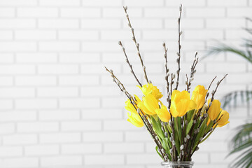Vase with pussy willow branches and tulip flowers near white brick wall, closeup