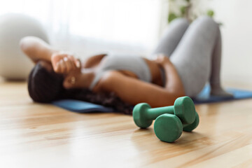 Tired African american woman working out in home livingroom gym with dumbbell in font view focus