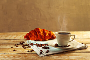 Coffee in a white ceramic mug. Croissant with coffee for breakfast. Coffee on an old wooden table. Coffee beans are scattered near the mug.
