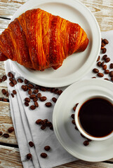 Coffee in a white ceramic mug. Croissant with coffee for breakfast. Coffee on an old wooden table. Coffee beans are scattered near the mug.